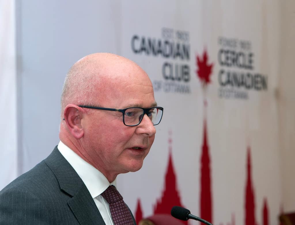 A speaker in glasses at the podium in front of the Club backdrop